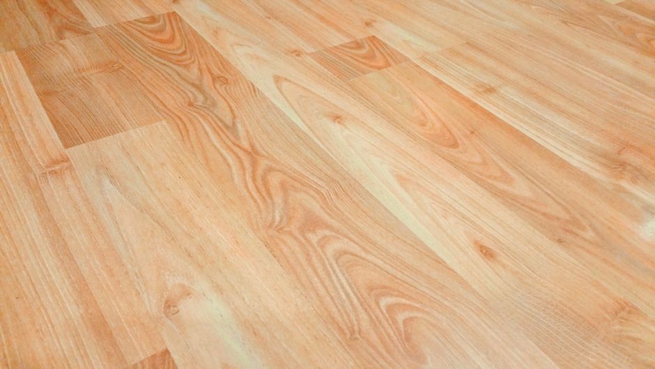 Close-up view of a wooden laminate flooring in a residential room, featuring a natural wood grain pattern with warm tones and subtle variations in color and texture. The surface appears clean and dry,