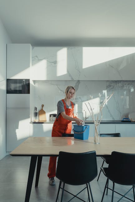 Two professional cleaners from House Cleaning Services, dressed in grey tops and bright orange overalls, are performing surface cleaning in a modern living room. The cleaner on the left is vacuuming t
