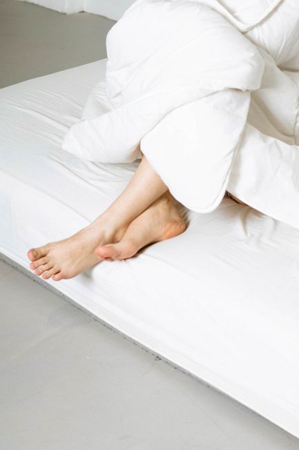 A professional cleaner from House Cleaning Services, dressed in a white protective suit, blue gloves, and a face mask, is deep cleaning a mattress at home. The cleaner is holding a handheld steam clea